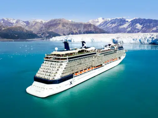 A large Celebrity cruise ship with multiple decks sailing through Hubbard Glacier, surrounded by icy blue waters and rugged mountainous terrain