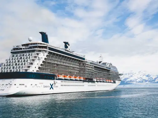 A large Celebrity cruise ship with multiple decks sailing through Hubbard Glacier, surrounded by icy blue waters and rugged mountainous terrain