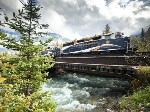 A scenic view of the Rocky Mountaineer train crossing a bridge over a rushing river. 