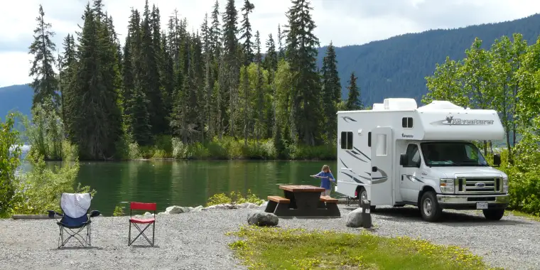 A motorhome at a campground in front of a tranquil lake setting with tall evergreen trees